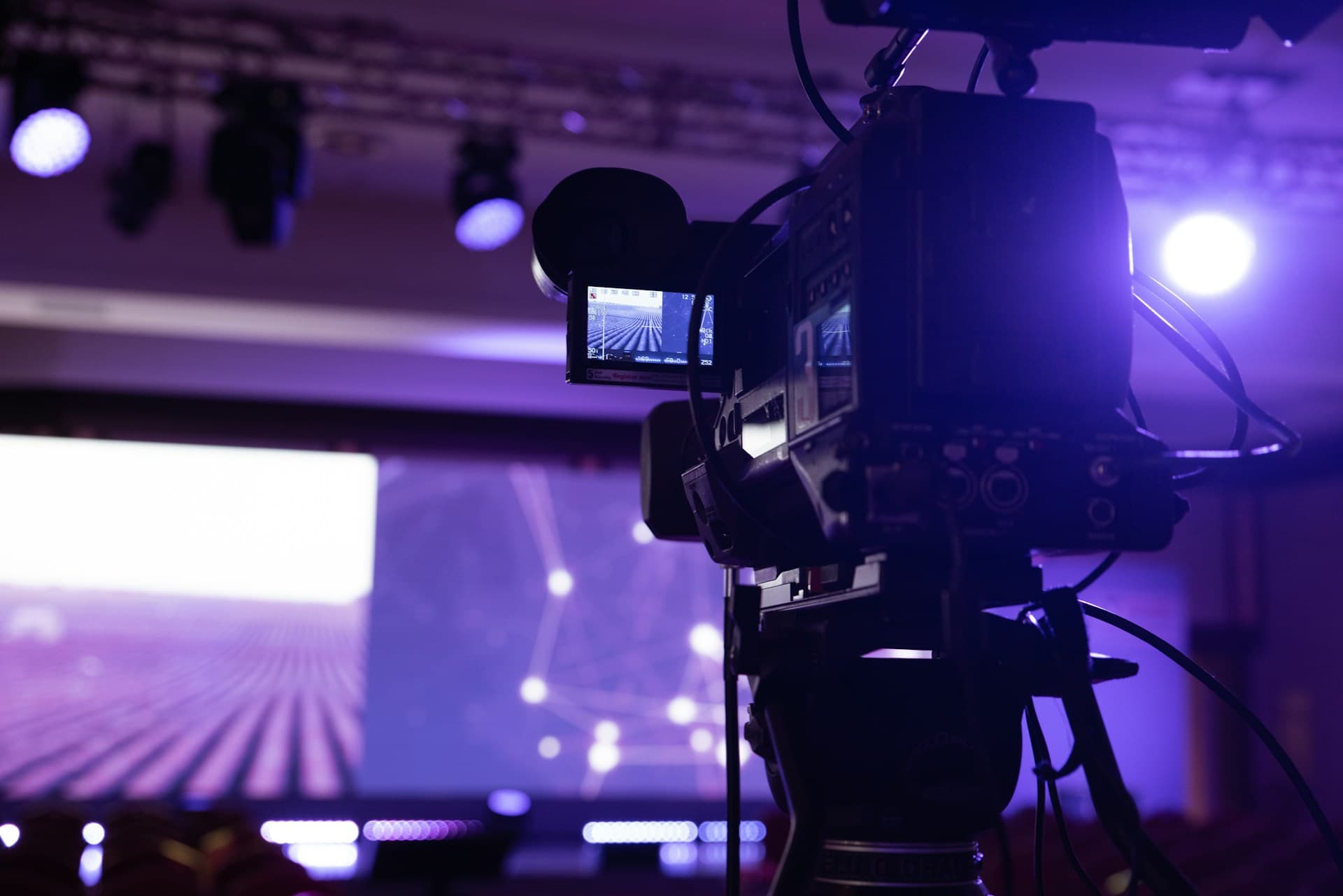 A broadcast camera on a tripod filming a lit stage